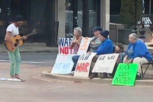 Photo of guitarist singing to people with signs protesting war.