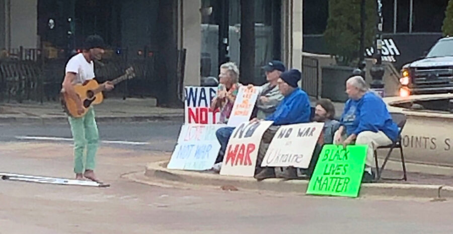 Photo of guitarist singing to people with signs protesting war.